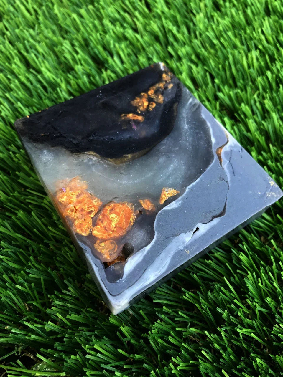 Handmade resin coaster with black and grey marble swirls and gold foil accents on green grass.